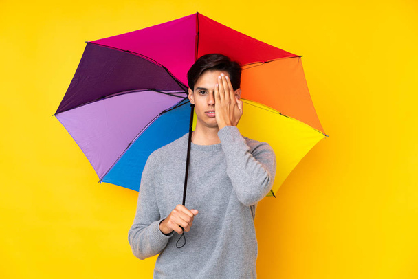 Man holding an umbrella over isolated yellow background covering a eye by hand - Foto, immagini