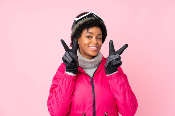 Mujer esquiadora afroamericana con gafas de snowboard sobre fondo rosa aislado sonriendo y mostrando signo de victoria
 - Foto, Imagen