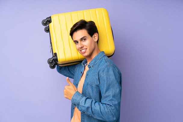 Man over isolated purple background in vacation with travel suitcase and with thumb up - Photo, image