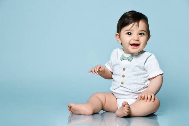 Little kid in white bodysuit as vest with bow-tie, barefoot. He smiling, sitting on the floor against blue background. Close up - Φωτογραφία, εικόνα