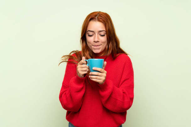 Adolescente rousse fille avec pull sur fond vert isolé tenant tasse chaude de café
 - Photo, image