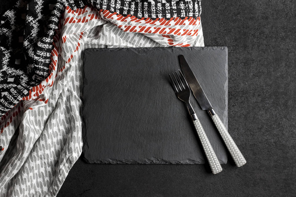 Placa de pizarra negra con tenedor y cuchillo sobre fondo negro y mantel. Configuración de la mesa. Plano, vista superior, espacio para copiar
 - Foto, imagen