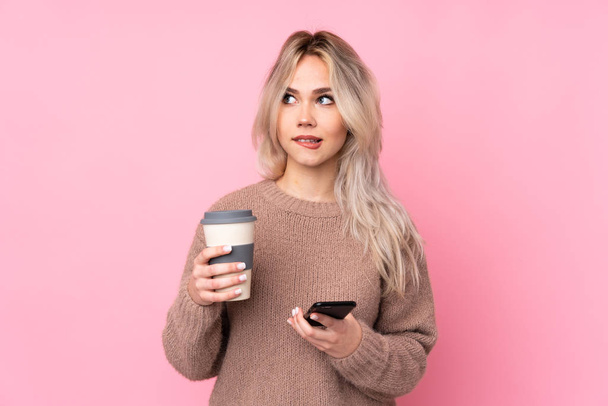 tiener blond meisje het dragen van een trui over geïsoleerde roze achtergrond houden koffie mee te nemen en een mobiele terwijl het denken iets - Foto, afbeelding