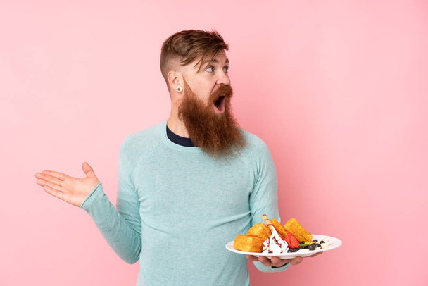 Redhead man with long beard holding waffles over isolated pink background with surprise facial expression - Фото, изображение