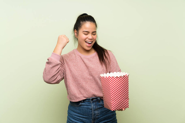 Young teenager Asian girl over isolated green background holding a bowl of popcorns - Photo, Image