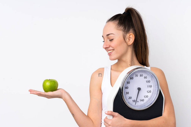 Młoda brunetka kobieta nad odizolowanym białym tle trzymając wagą podczas patrząc jabłko - Zdjęcie, obraz