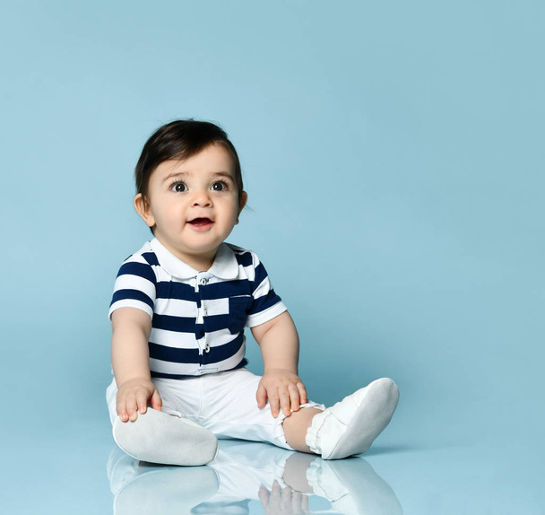 Baby boy in striped t-shirt, white pants and booties. He is smiling, sitting on floor against blue studio background. Close up - Valokuva, kuva