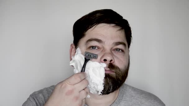 Een man scheert een dikke, grote baard af met een scheermes. Een man met baard scheert zich met scheerschuim. Gezichtsbehandeling. Portret - Video