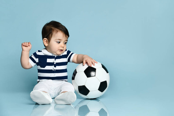 Toddler in striped t-shirt, white pants and booties. He is looking up, sitting on floor against blue background. Close up - Fotoğraf, Görsel