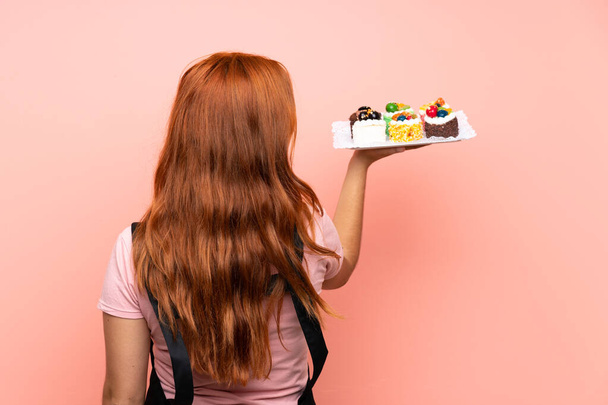 Adolescente ruiva menina segurando lotes de diferentes mini-bolos sobre isolado fundo rosa na posição de trás
 - Foto, Imagem
