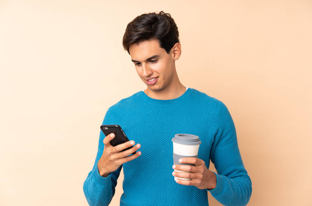 Man over isolated background holding coffee to take away and a mobile - Photo, Image