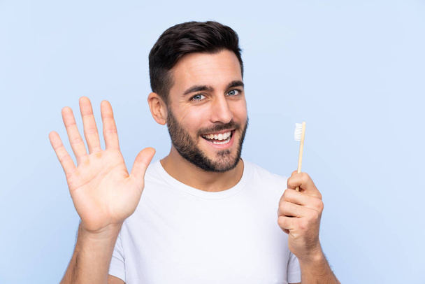 Молодий красивий чоловік з бородою чистить зуби над ізольованим фоном соління рукою з щасливим виразом
 - Фото, зображення