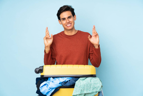 Мужчина с чемоданом, полным одежды на изолированном синем фоне с скрещенными пальцами
 - Фото, изображение