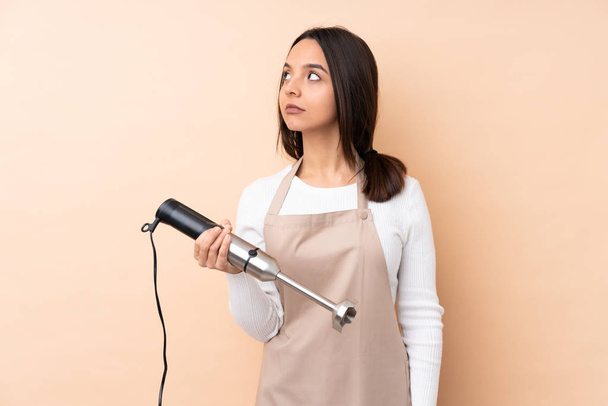 Young brunette girl using hand blender over isolated background making doubts gesture looking side - Foto, afbeelding