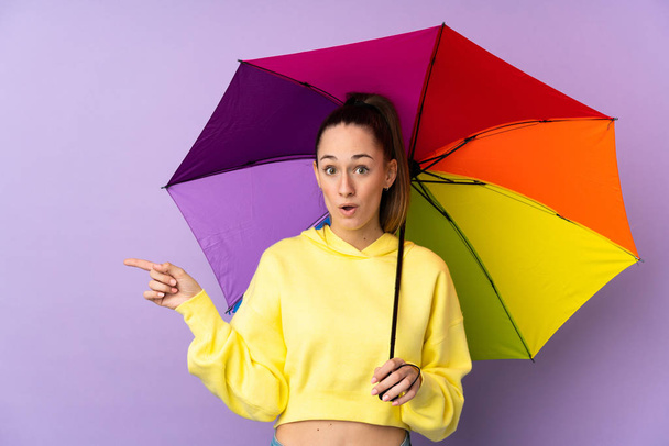 Joven morena sosteniendo un paraguas sobre aislada pared púrpura sorprendida y señalando lado
 - Foto, imagen