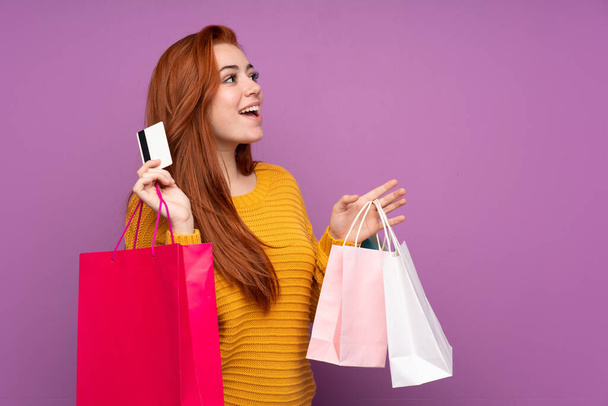 Piros hajú tinédzser lány felett elszigetelt lila háttér gazdaság bevásárló táskák és egy hitelkártya - Fotó, kép