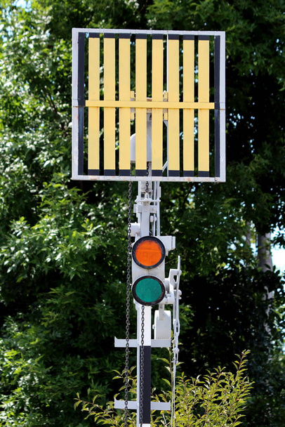 Feux de circulation rouges et verts au bord de la voie ferrée sous le panneau de signalisation monté sur poteau métallique à la gare locale avec des arbres denses en arrière-plan
 - Photo, image