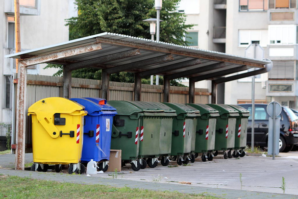 Reihe von verschiedenen großen Plastikmüllcontainern zur ökologischen Mülltrennung zum Recyceln unter dem Schutz von Metalldachziegeln zwischen alten hohen Wohnhäusern - Foto, Bild
