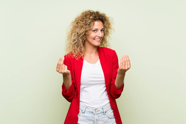 Young blonde woman with curly hair over isolated green background making money gesture - Fotografie, Obrázek