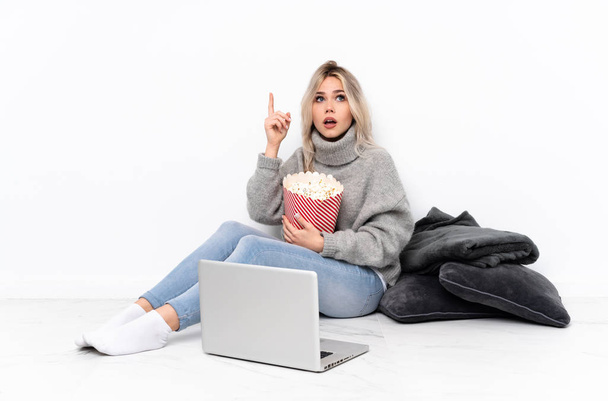 Teenager ξανθιά κοπέλα τρώει ποπ κορν, ενώ βλέποντας μια ταινία στο φορητό υπολογιστή με σκοπό να πραγματοποιήσει τη λύση, ενώ σηκώνοντας ένα δάχτυλο προς τα πάνω - Φωτογραφία, εικόνα