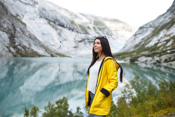 The girl tourist in a yellow jacket posing on the lake in Norway. Active woman relaxing near the lake against the backdrop of the mountains in the Norway. Travelling, lifestyle, adventure, wild nature - Photo, Image
