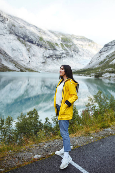 The girl tourist in a yellow jacket posing on the lake in Norway. Active woman relaxing near the lake against the backdrop of the mountains in the Norway. Travelling, lifestyle, adventure, wild nature - Foto, immagini