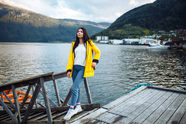 The girl tourist in a yellow jacket posing by the lake in Norway. Active woman relaxing near the boat by the lake against the backdrop of the mountains in the Norway. Travelling, lifestyle, adventure. - Foto, imagen
