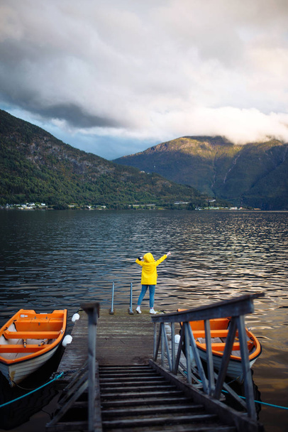 The girl tourist in a yellow jacket posing by the lake in Norway. Active woman relaxing near the boat by the lake against the backdrop of the mountains in the Norway. Travelling, lifestyle, adventure. - Photo, image