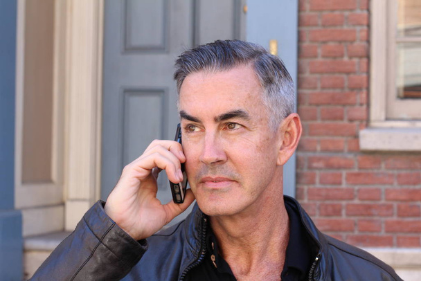 close-up portrait of handsome mature man in leather jacket talking by phone on street - Foto, imagen