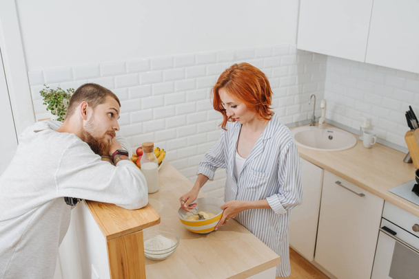 Wife cooking, whisking mixture in a bowl, while her husband entertains her - Valokuva, kuva
