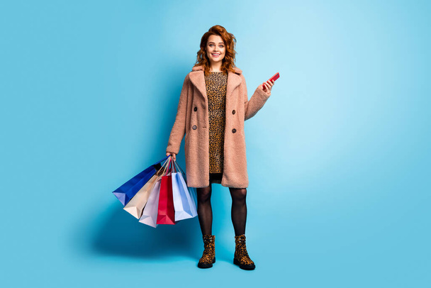 Full size photo of positive feminine lady tourist walk fun use smartphone to pay easy shopping hold many bags wear season coat dress tights cotton boots isolated over blue color background - Fotografie, Obrázek