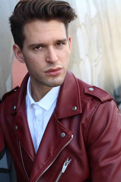 Bad boy concept. Portrait of brutal young man with short hair wearing red leather jacket, jeans and posing over urban background. Hand in pockets. Hipster style. Copy-space. Outdoor shot - Φωτογραφία, εικόνα