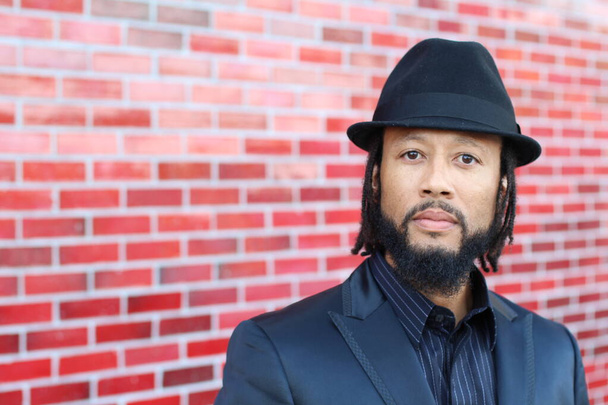 close-up portrait of handsome young man in black suit with hat and dreadlocks in front of red brick wall - Foto, imagen