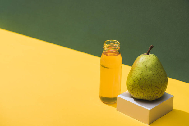 fresh juice in bottle near pear and white cube on green and yellow background - Fotografie, Obrázek