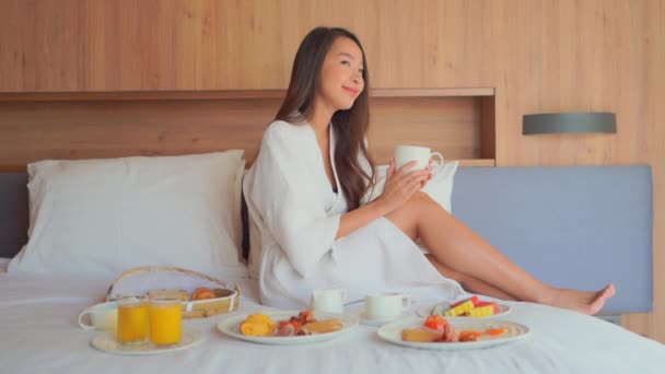 Filmaufnahmen der schönen asiatischen Frau, die Kaffee trinkt, während sie zu Hause im Bett frühstückt - Filmmaterial, Video