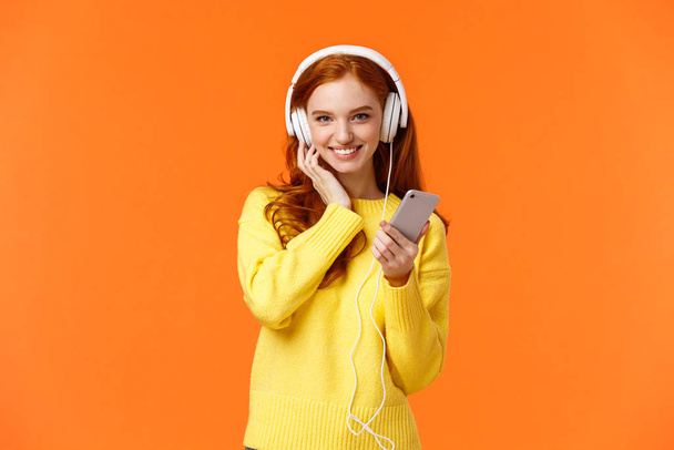 Sassy good-looking redhead female in yellow sweater, listen music white headphones, touch earphones and holding smartphone, listen music, enjoy good earbuds quality, smiling, orange background - Fotó, kép
