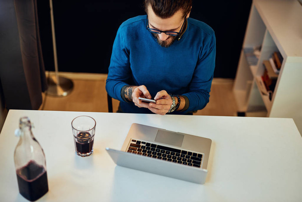 Aantrekkelijke blanke bebaarde ondernemer zit in zijn kantoor, met behulp van een smartphone en het nemen van een pauze van het werk. - Foto, afbeelding