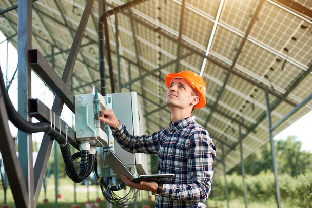 Male engineer in helmet and with laptop pushing button at the electrical equipment on solar power station, looking up. Science technology. Solar panels. Global warming. Green energy. - Photo, image