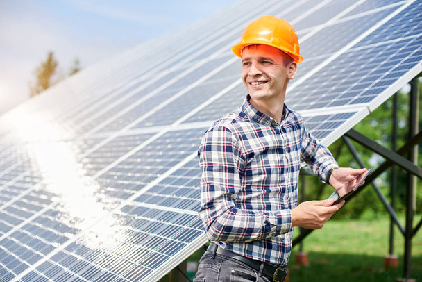 Ευτυχισμένος τύπος σε ένα κράνος με μια ταμπλέτα στα χέρια του κοιτάζοντας μακριά κοντά στο σταθμό ηλιακών κυττάρων με μια αντανάκλαση του ήλιου σε αυτά. Οικοδομή. Παραγωγή πράσινης οικολογικής ενέργειας - Φωτογραφία, εικόνα