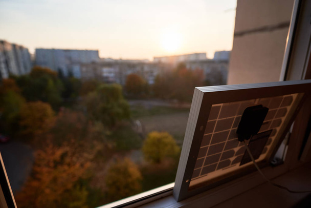 高層住宅の窓の上に立つ太陽電池の中からの眺め。窓の外では、秋の日に街の睡眠エリアで日没. - 写真・画像