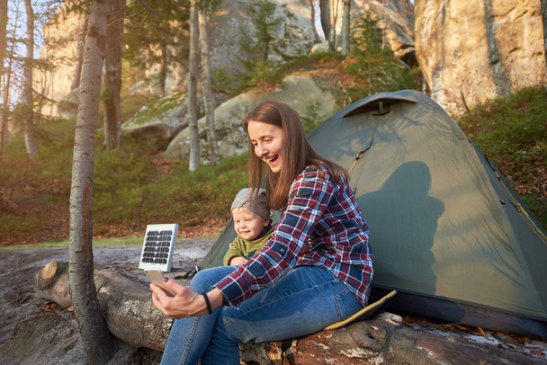 母親と赤ちゃんは、森の崖のふもとにソーラーパネルを備えた観光テントを背景に、携帯電話で自撮りをします。太陽の下での家族はカメラで穏やかな笑顔を見せる - 写真・画像