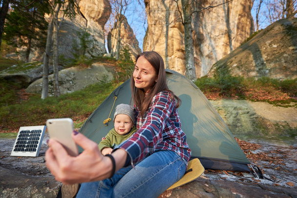 母親と息子は、森の崖のふもとにソーラーパネルを持つ観光テントの背景にある携帯電話で自撮りをします。優しい笑顔でカメラを見ている若い家族 - 写真・画像