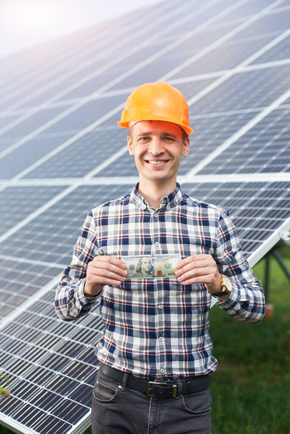 Das Porträt eines glücklichen Mannes mit einem Hundert-Dollar-Schein in der Hand zeigt im Hintergrund einer Solarstation ein Schild, wie es ihm gefällt. Geschäfts- und Generationenkonzept. grüne ökologische Stromerzeugung. - Foto, Bild
