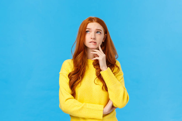 Perplexed, worried and concerned cute redhead student college girl trying choose university, thinking future, looking pensive upper left corner, bite lip and frowning, thoughtful over blue background - Photo, Image
