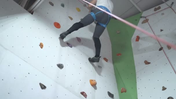 Sporty young man climbing indoors with belay, descending from top - Footage, Video