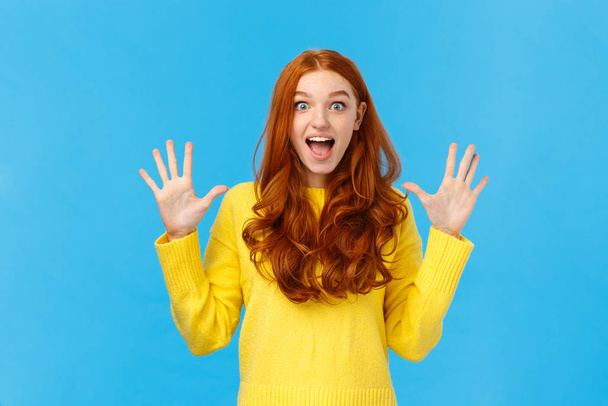 Excited, overwhelmed happy redhead cute girl in yellow sweater, screaming from joy telling about christmas gifts, showing ten fingers, order dozen products, standing joyful blue background - Foto, immagini