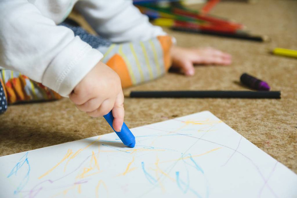 A closeup shot of a baby drawing with a blue crayon sitting on the floor - Fotó, kép