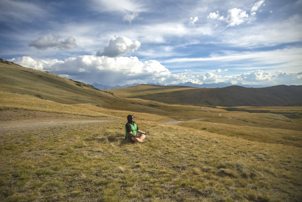 Joven turista relajado en las montañas, lejos de la vida de la ciudad y reuniones de negocios. Hombre solitario con capucha verde, sentado en la cima de la colina de la montaña bajo el cielo nublado dramático en la hierba alta y disfrutando de tranquilidad y soledad. Montaña Altai
 - Foto, imagen