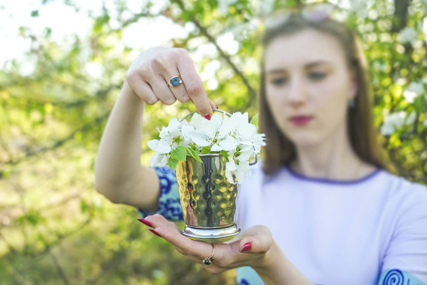 A bouquet of spring flowers in the hands of a girl on a background of blue dress. Blooming apple tree in the park in spring.  branches of apple tree standing in iron metal bucket. blurred face - Foto, afbeelding