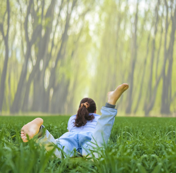 barefoot young woman lying in fresh green grass and reading a book. back view.woman lying on spring green grass. summer time season. unrecognizable person. springtime in park. - Photo, image